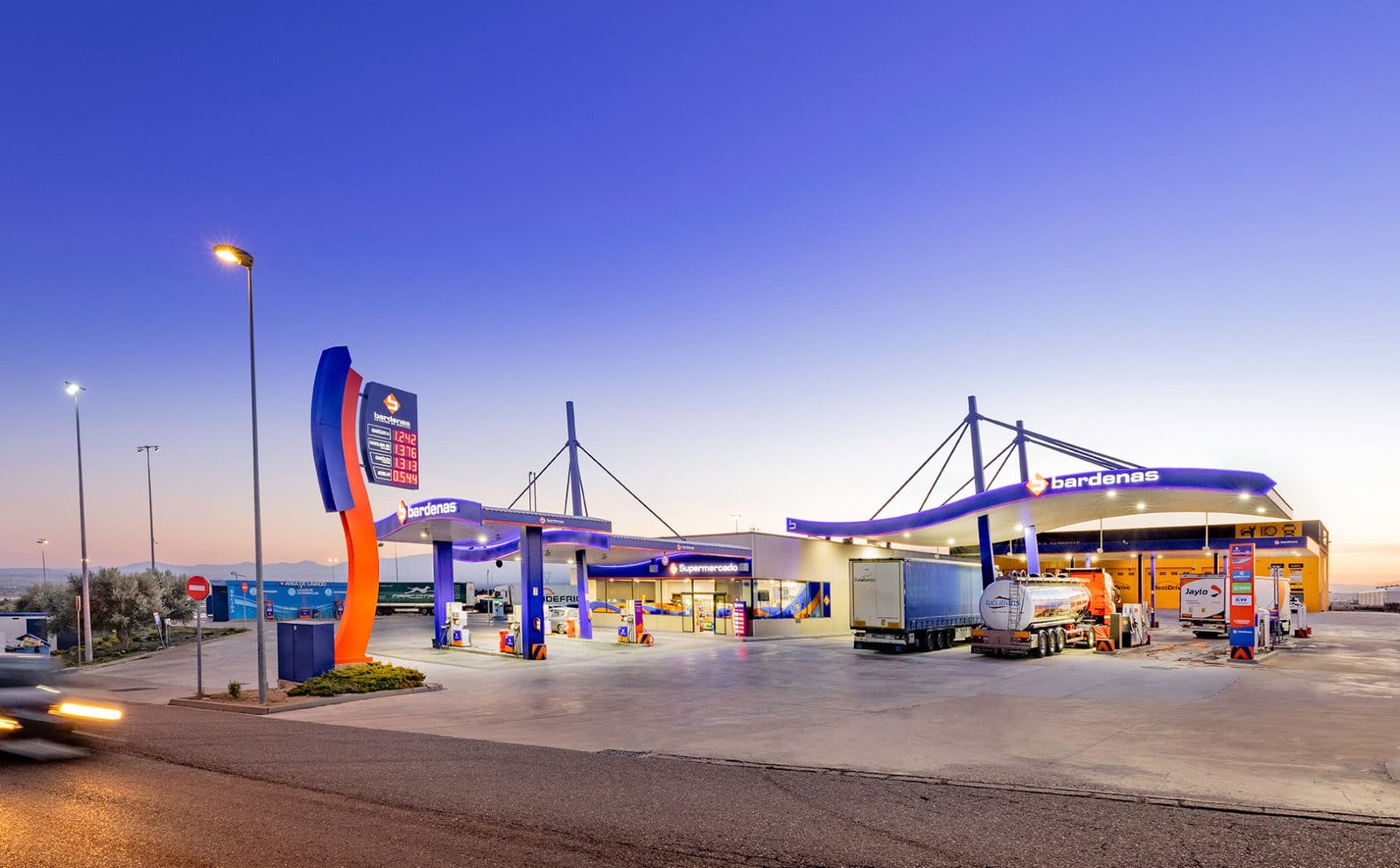 E.S. Bardenas al anochecer: tótem de precios, marquesinas iluminadas en azul y naranja, supermercado y camiones repostando